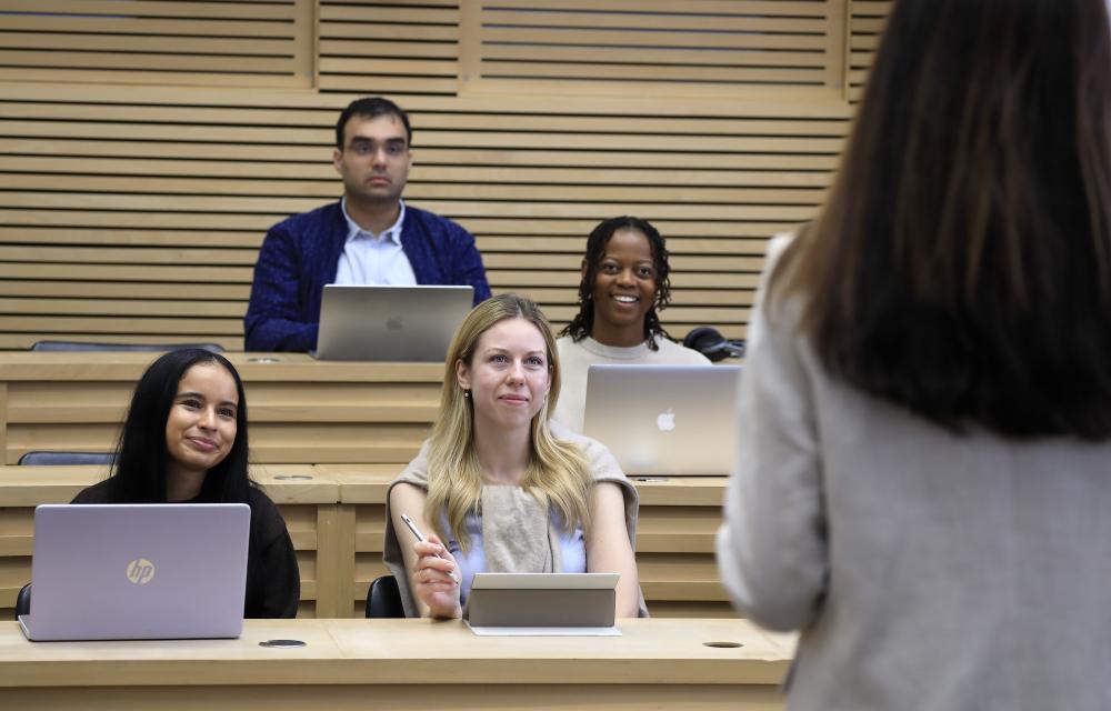 Graduate students in the Cube smiling whilst listening to a lecture. They have laptops on the desks in front of them.