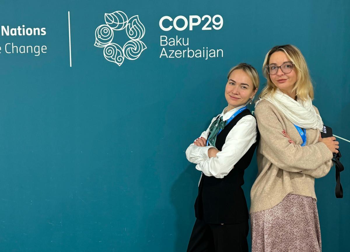 2 women- back to back in front of COP29 logo