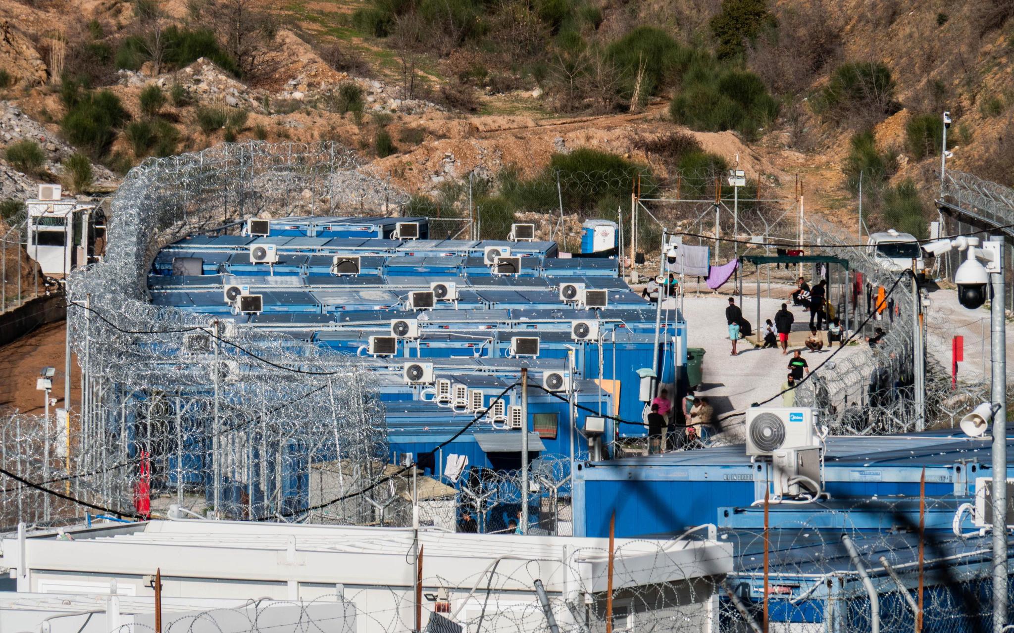 Blue containers on a hill, with air conditioner units and people walking around them. Extensive barbed wire encloses the units
