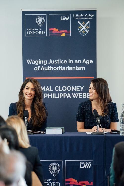 Amal Clooney and Philippa Webb sitting at a table with an oxford blue pop-up banner behind them.
