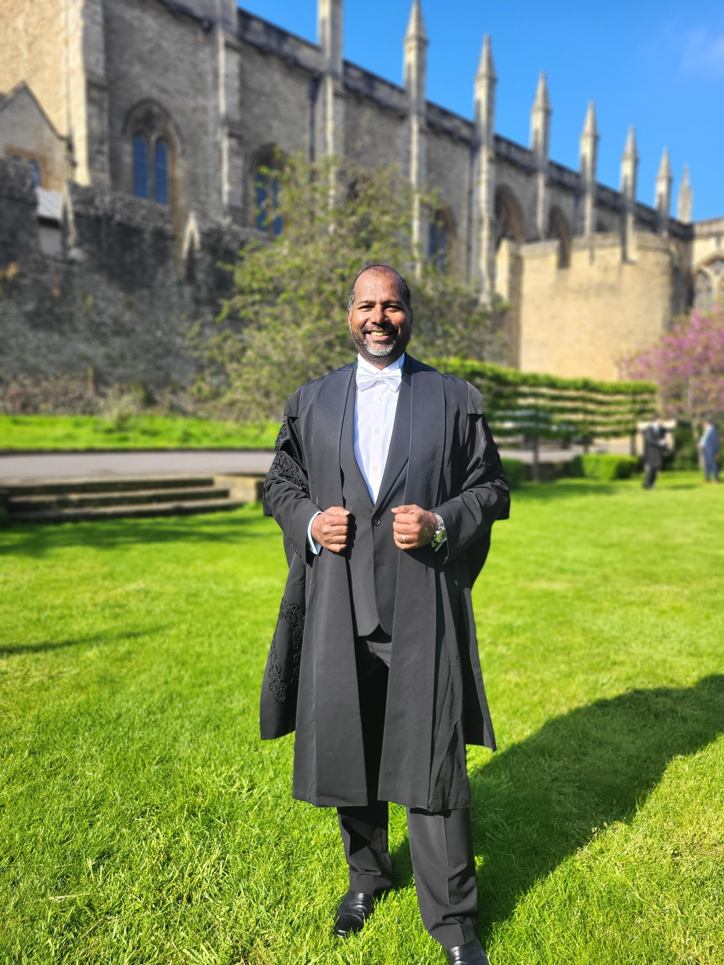 Man dressed in sub-fusc standing in front of Christchurch college on a sunny day.