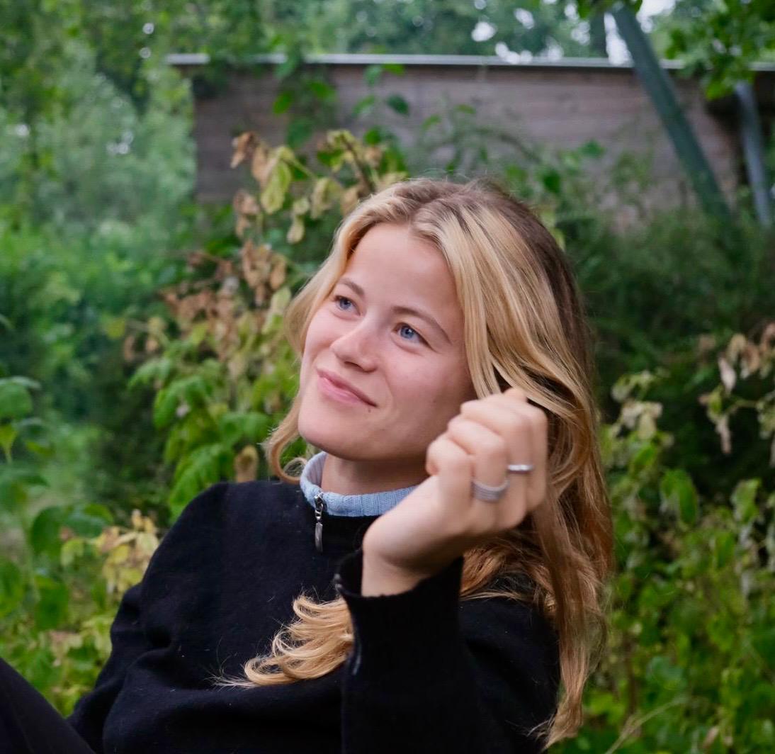 A woman with dyed blonde and brown hair smiles at something off camera with her hand raised near her face. She wears a black zip up fleece and is backdropped by greenery