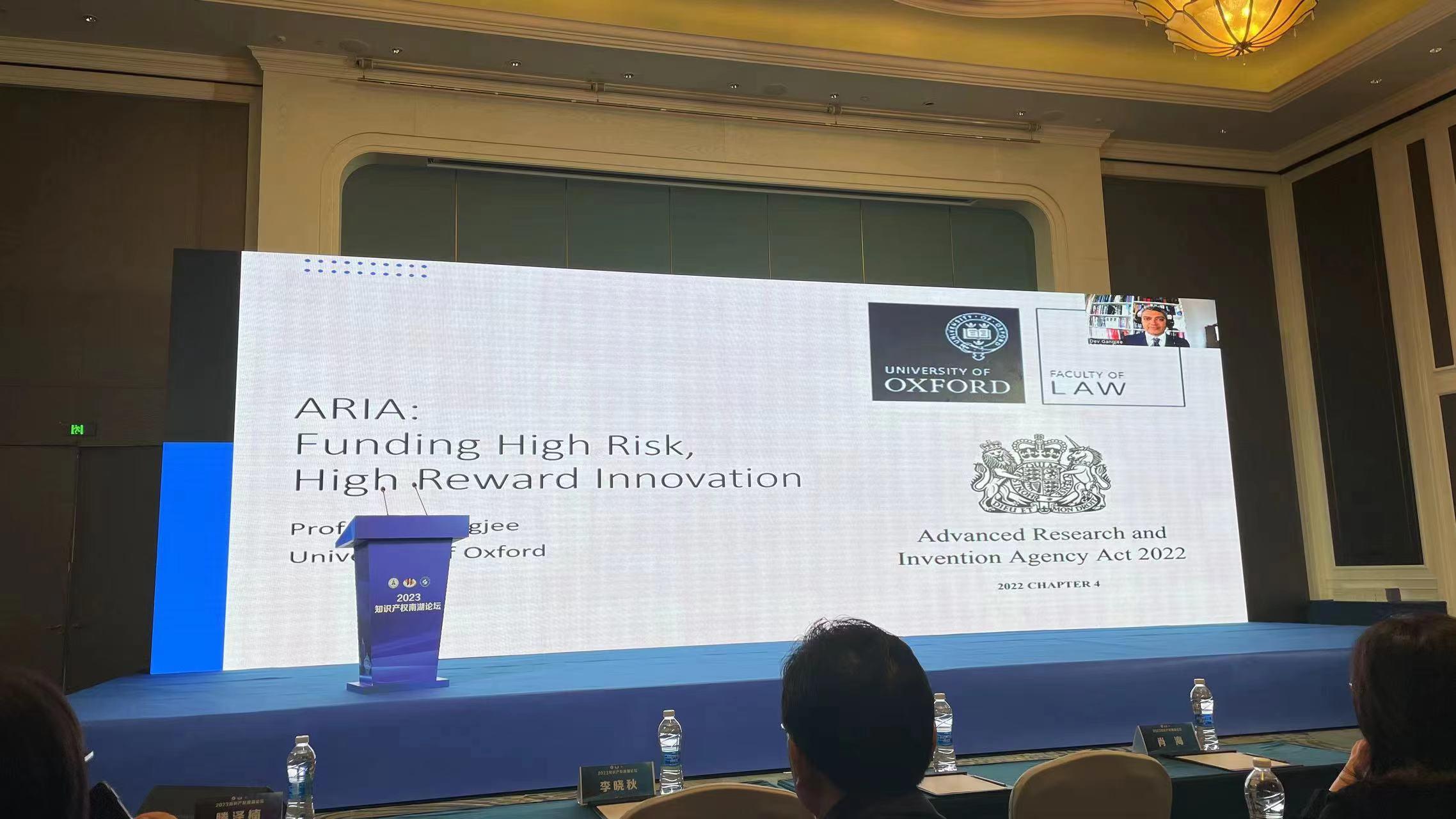 A large screen showing the title of the talk, with Dev Gangjee appearing remotely in the top right hand corner