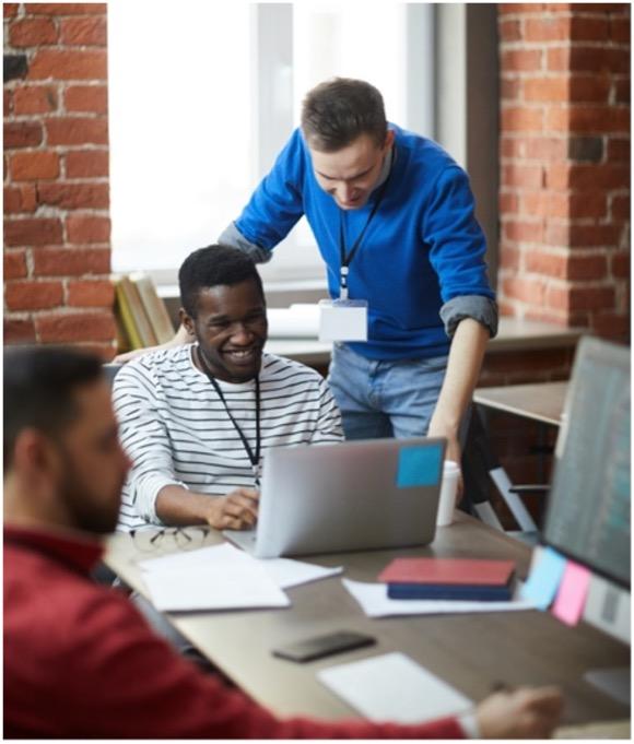 A man looking over another man's shoulder at a computer in an office setting.
