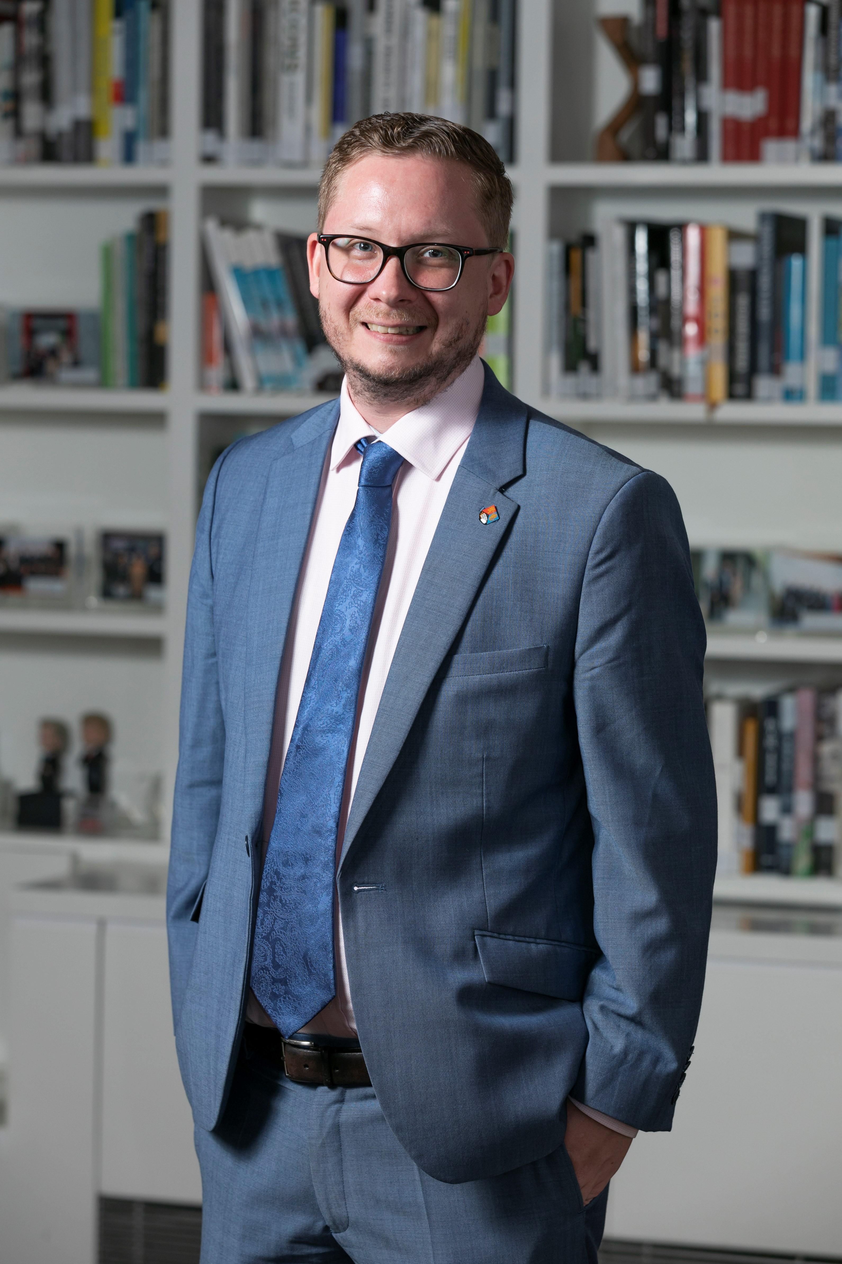 Craig Edwards - white man in a blue suit and tie standing infront of bookshelves
