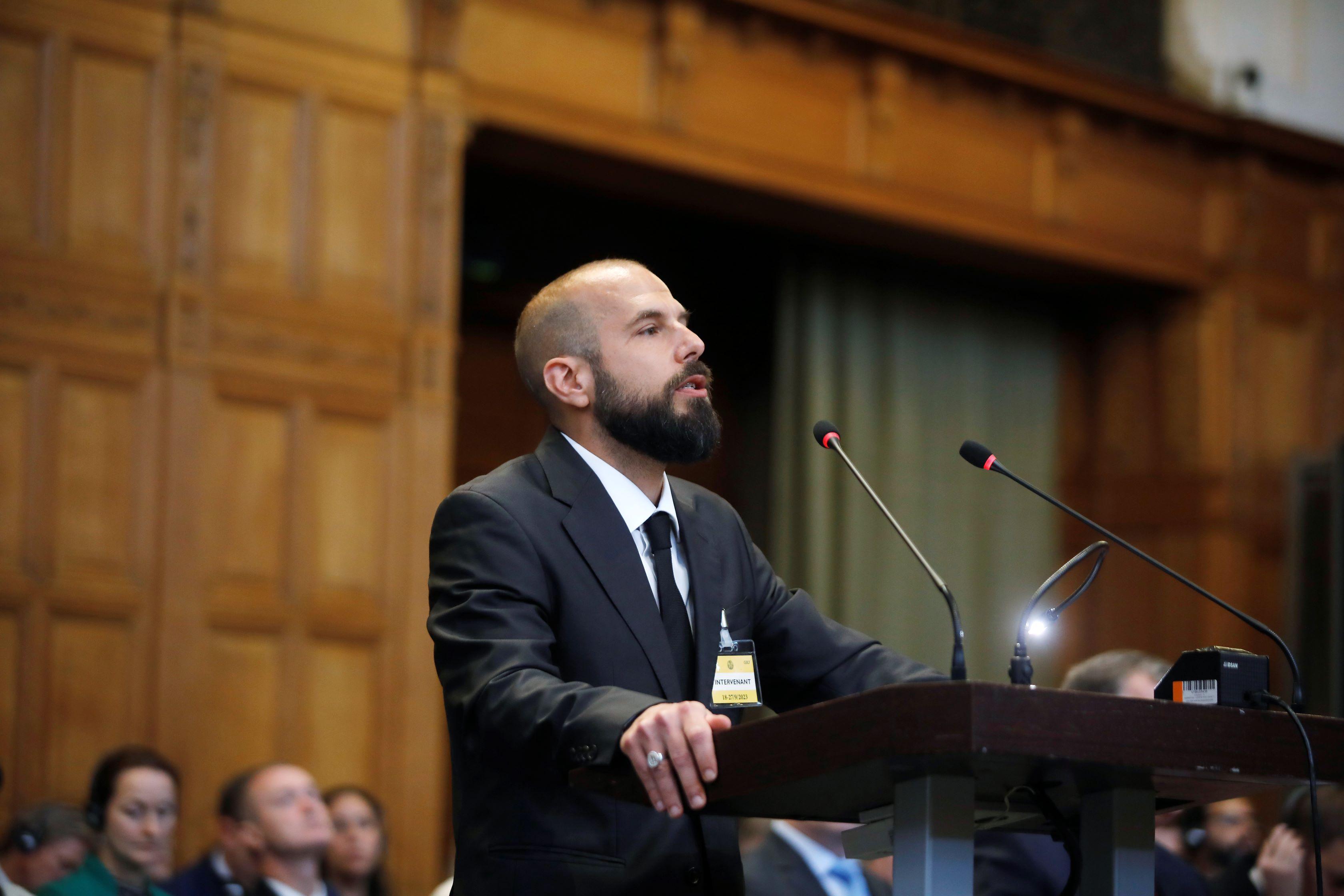 A bald white man with a beard standing at a lecturn in a wood panelled room