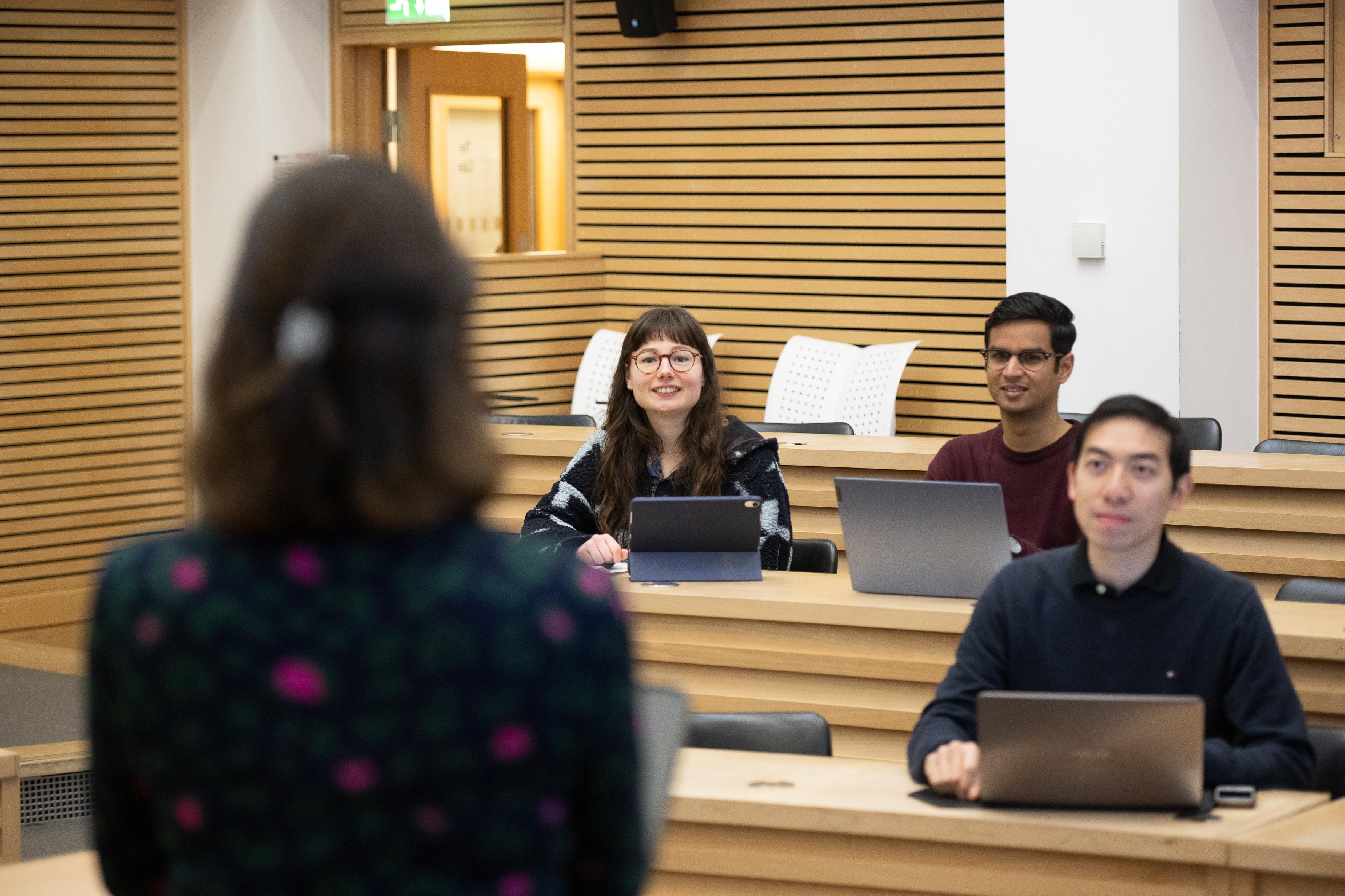 three students sitting in a lecture theatre, facing a lecturer who has her back to the camera.