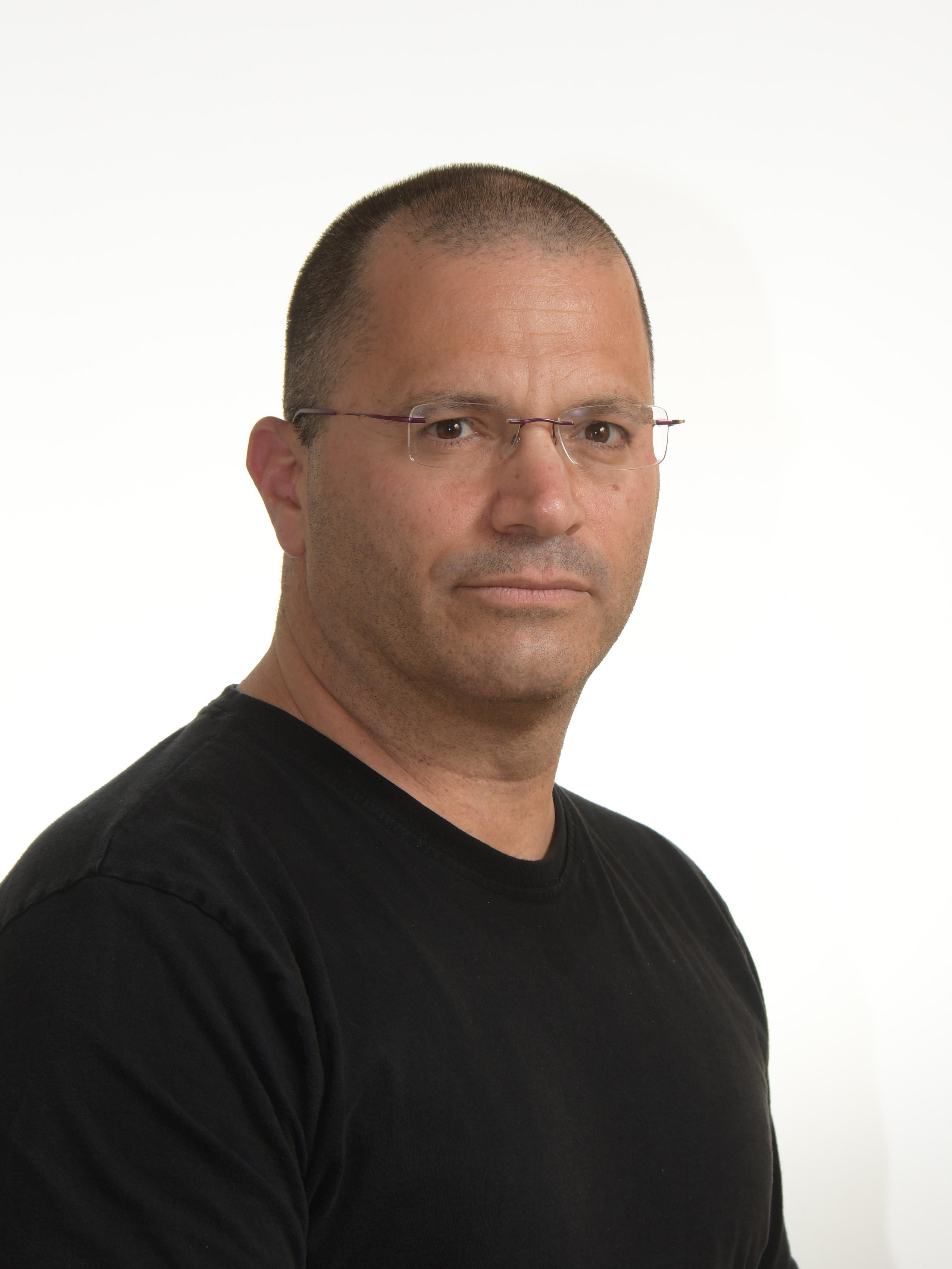 A man facing the camera with very short hair, wearing glasses and a black top