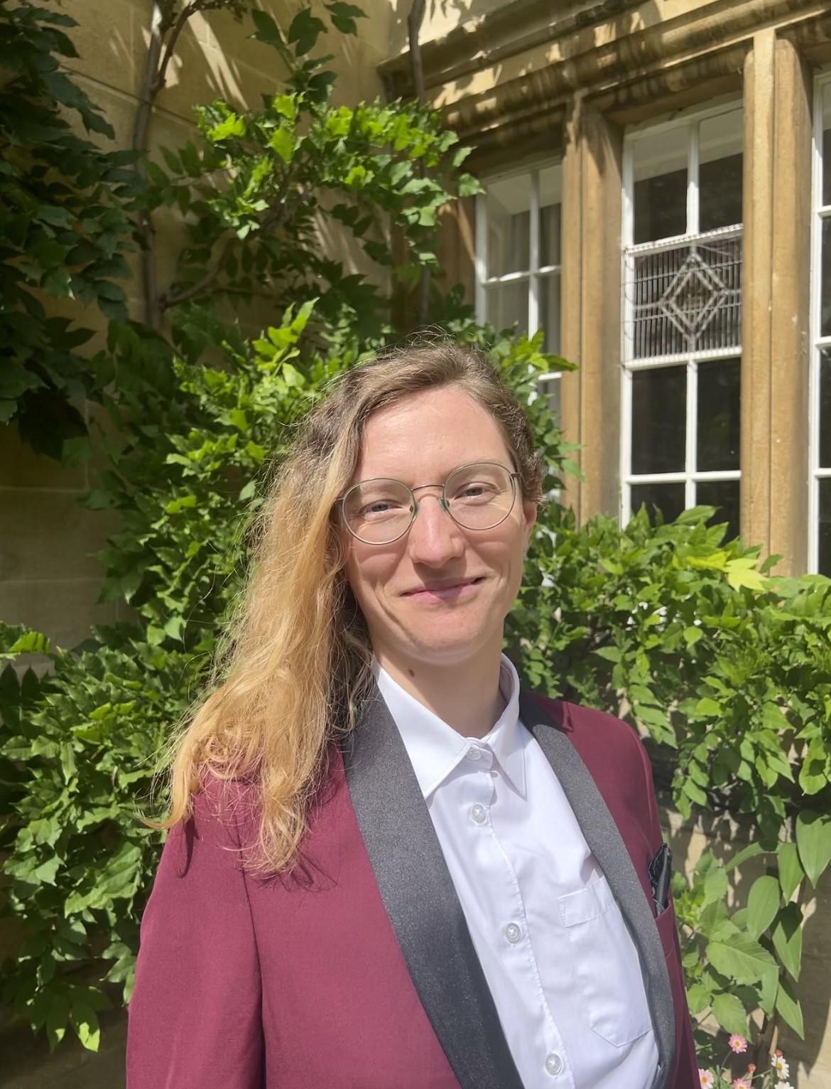 Profile picture of Sarah Ourednickova, young blonde woman with glasses in a white shirt and burgundy suit, against a college building covered in green plants
