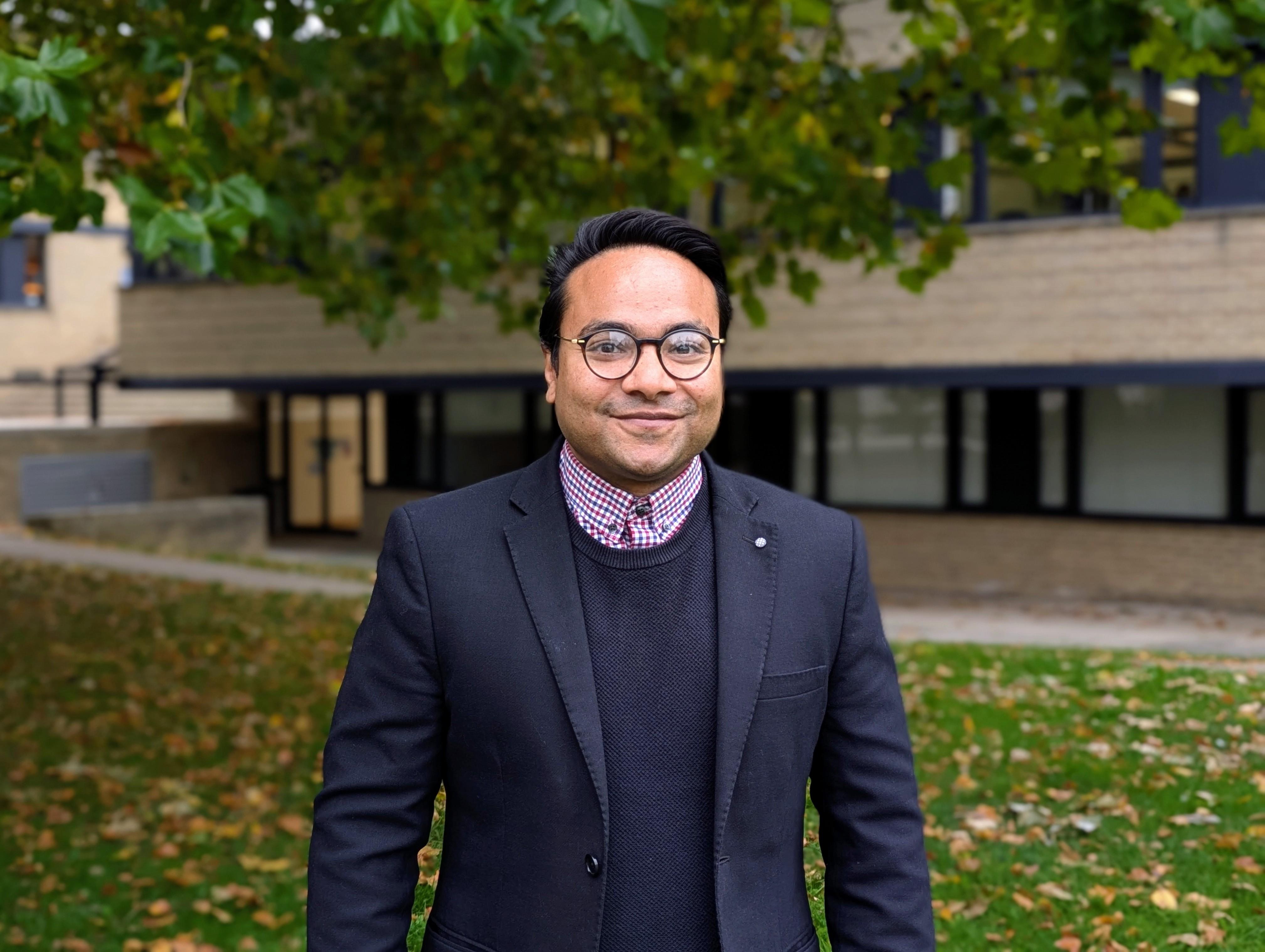 Asian man with glasses standing in front of tree outside St Cross Building smiling.