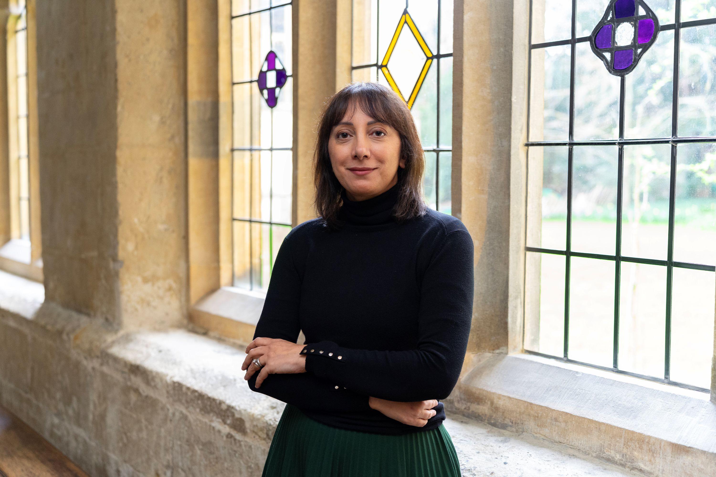  A woman with dark hair and olive skin wearing a black polo neck stands in front of a stained glass window