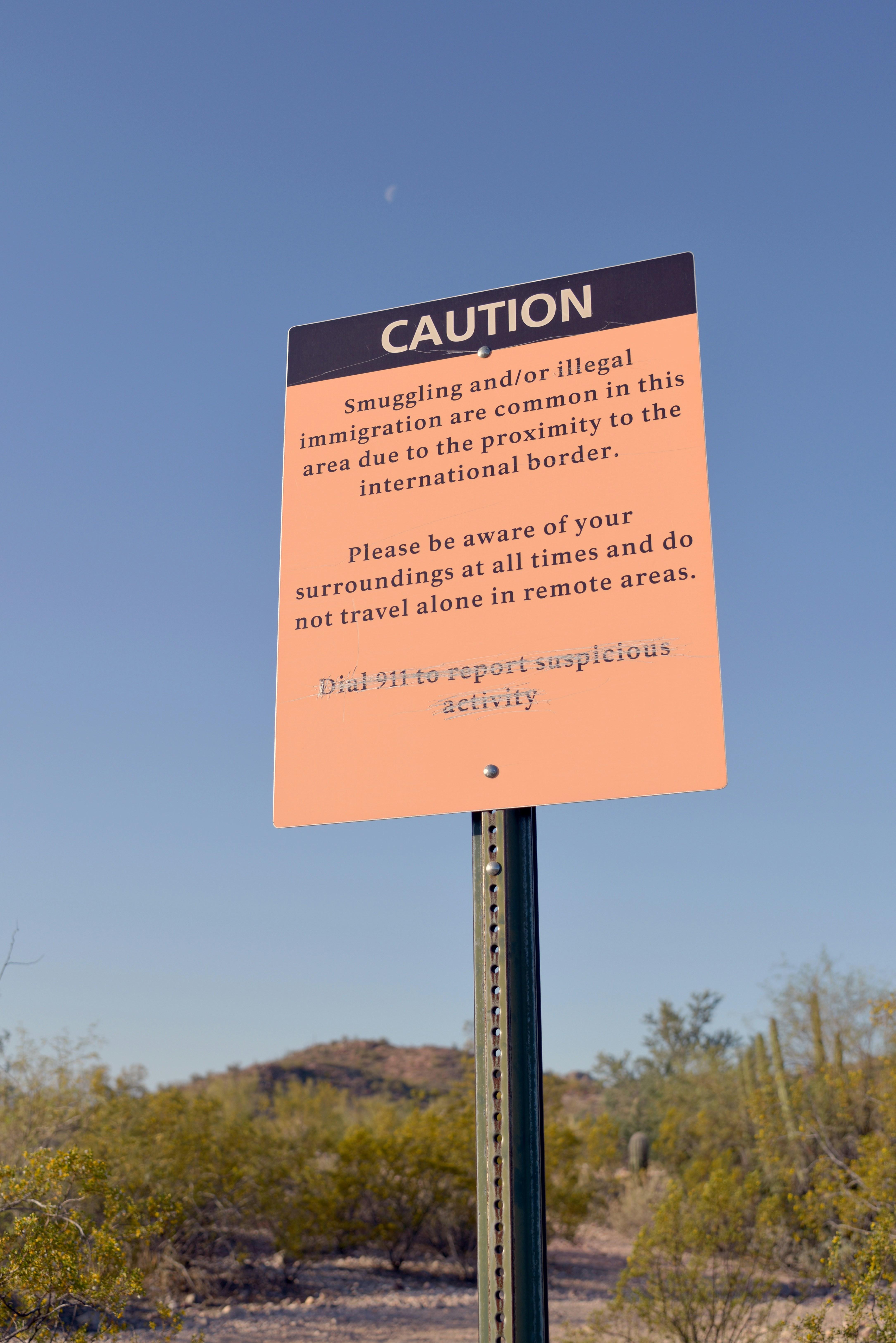 An orange caution sign stands against a backdrop of blue sky and desert. It reads: Caution. Smuggling and/ or illegal immigration are common in this area due to the proximity to the international border. Please be aware of your surroundings at all times and do not travel alone in remote areas. Graffiti paint has crossed out the next line: Dial 911 to report suspicious activity