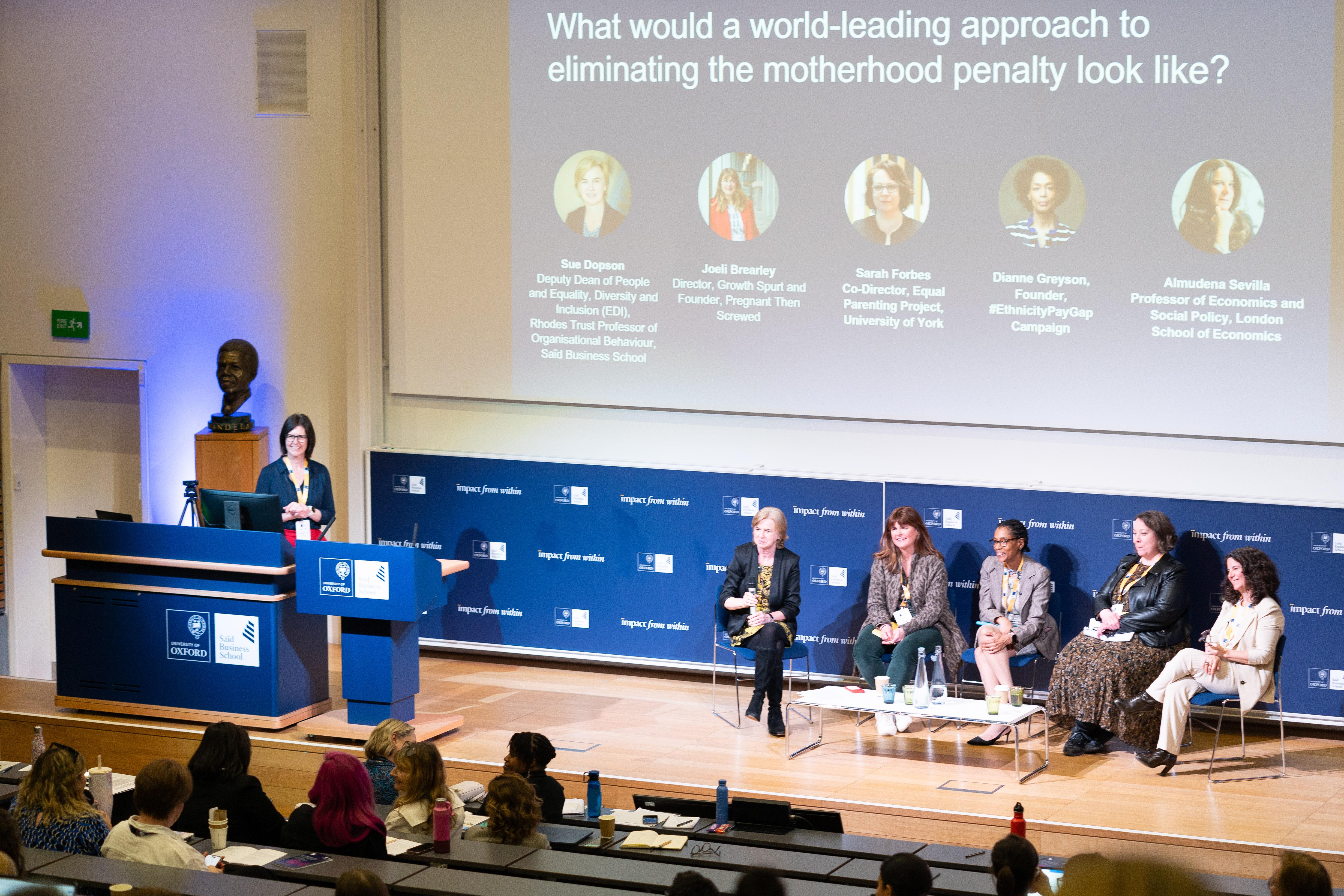 Presenter and panel on a stage with a blue background behind them.