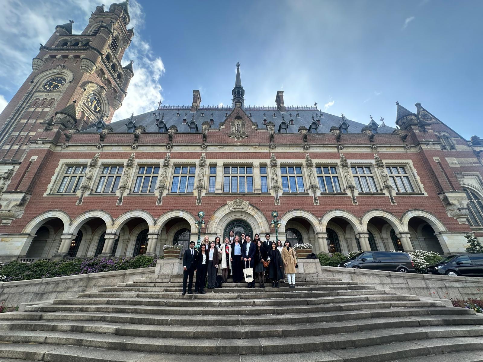 A group of 15 students standing on steps in front of the International Courts of Justice, a beautiful old building with a tower and colonades.