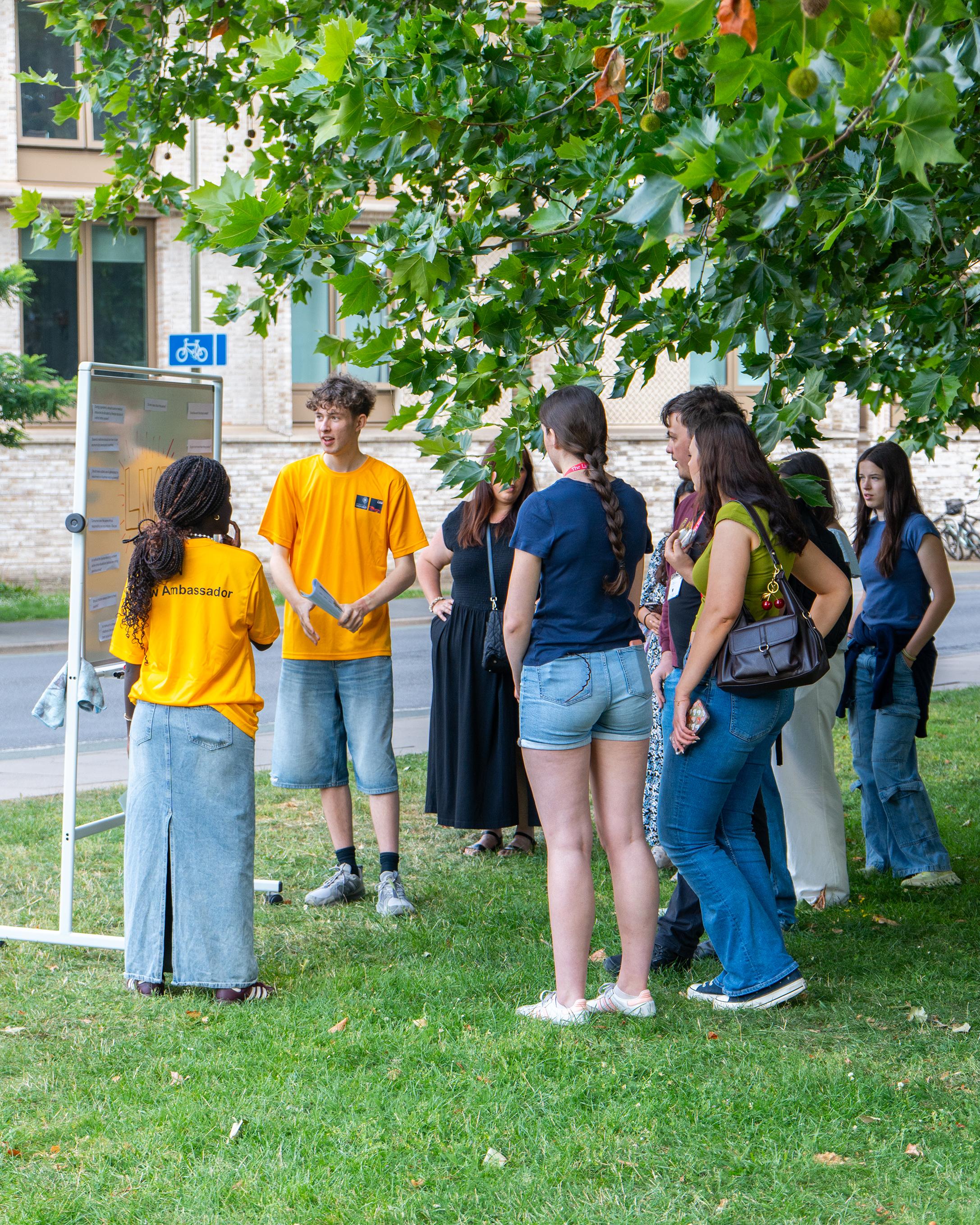 Two student ambassadors presenting to a crowd at an Open Day using a whiteboard on the grass outside the St Cross Building