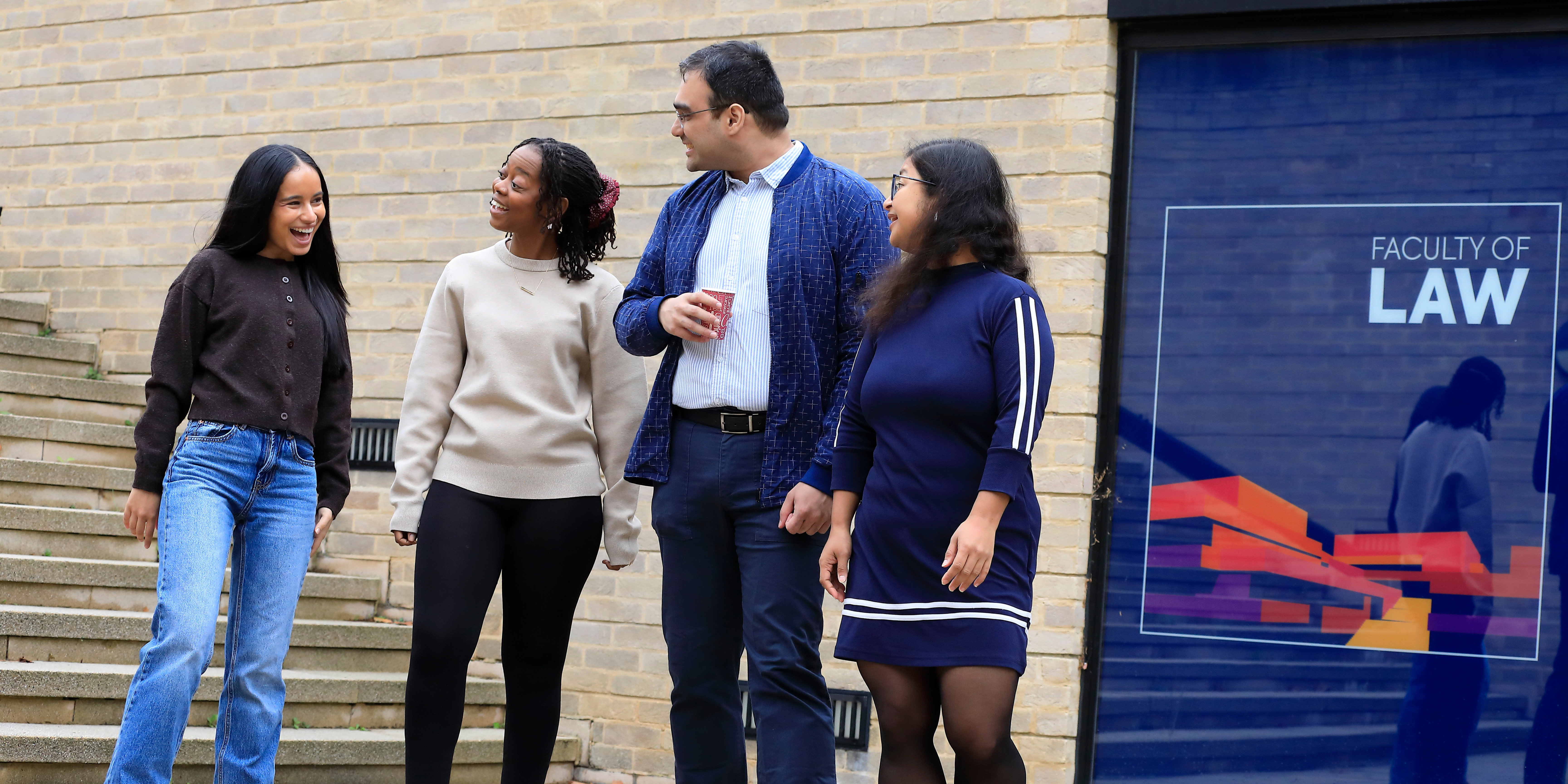 Four students walking and talking outside the St Cross Building with the Faculty of Law logo on a window in the background